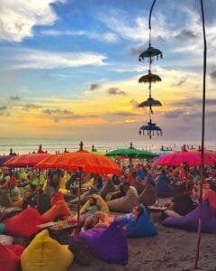 Colorful bean bags and umbrellas at a vibrant beach bar during sunset, a popular attraction and one of the must visit places in Seminyak, Bali, offering stunning ocean views and a lively atmosphere.