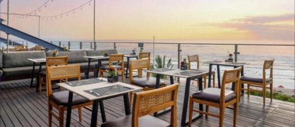 Outdoor dining setup at one of the stunning seaside restaurants in Bali, featuring wooden tables and chairs overlooking the beach with breathtaking ocean views during sunset.