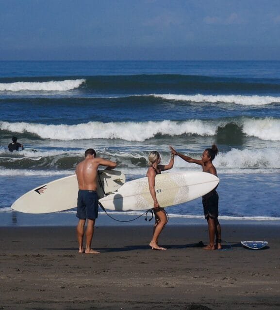 "Surfers at Kedungu Beach Bali celebrating a successful surf session with a high-five on the black sand shore, while waves roll in the background, creating a perfect setting for a surf adventure in Bali."