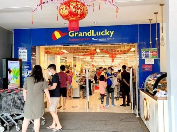 Entrance of Grand Lucky Bali supermarket decorated with red Chinese lanterns, shoppers entering and exiting, and trolleys lined up outside on Sunset Road